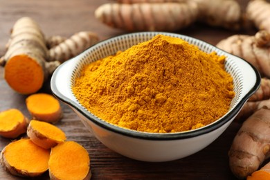 Photo of Bowl with aromatic turmeric powder and cut roots on wooden table, closeup