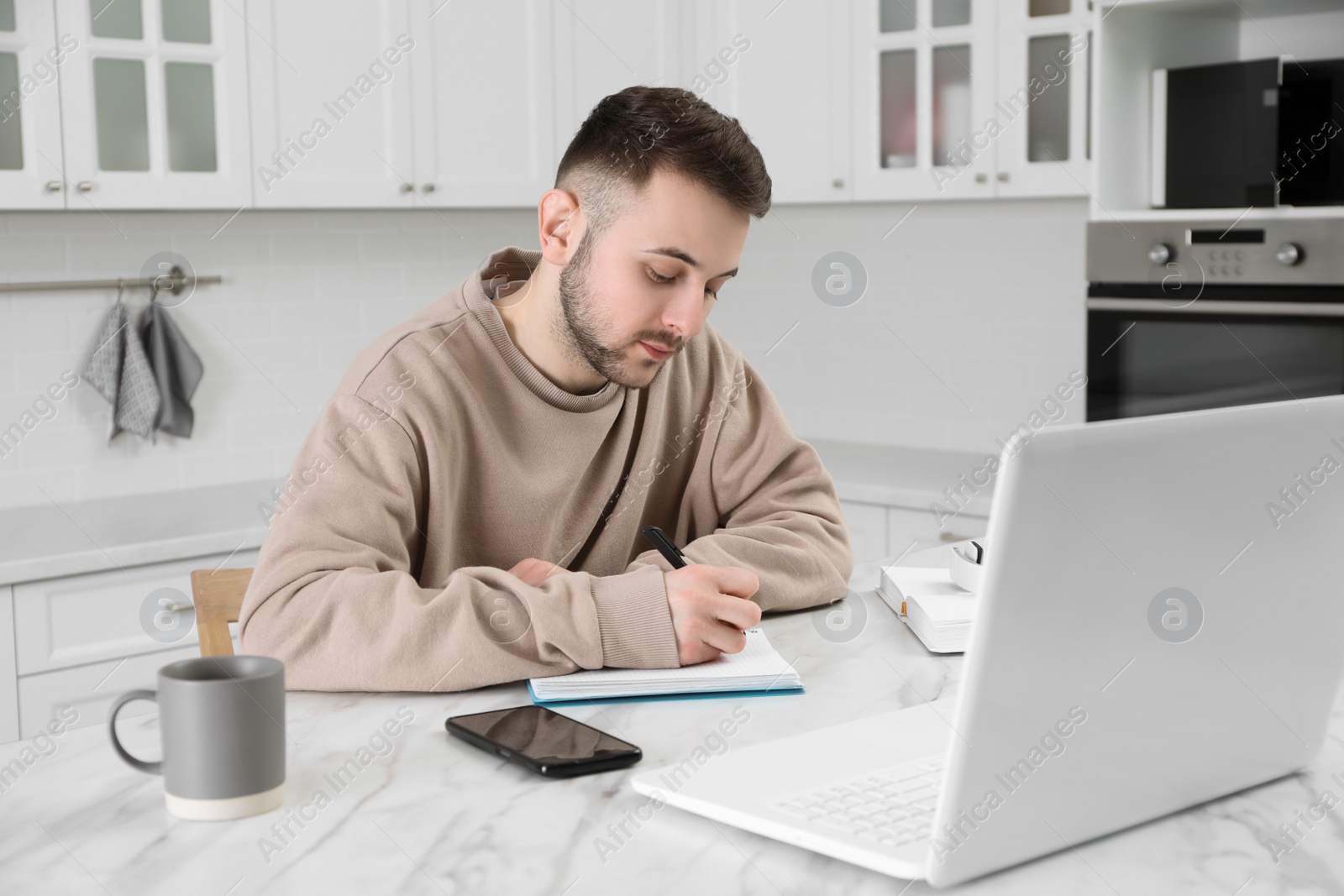 Photo of Young man using modern laptop for studying in kitchen. Distance learning