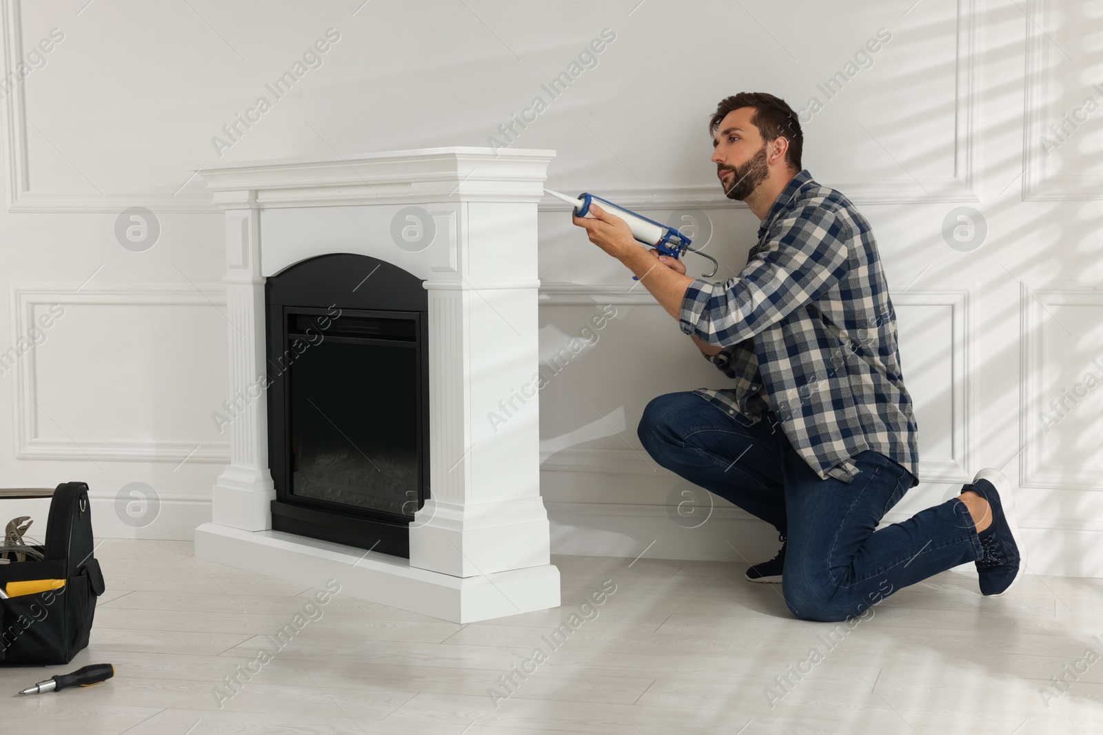 Photo of Man sealing electric fireplace with caulk near white wall in room