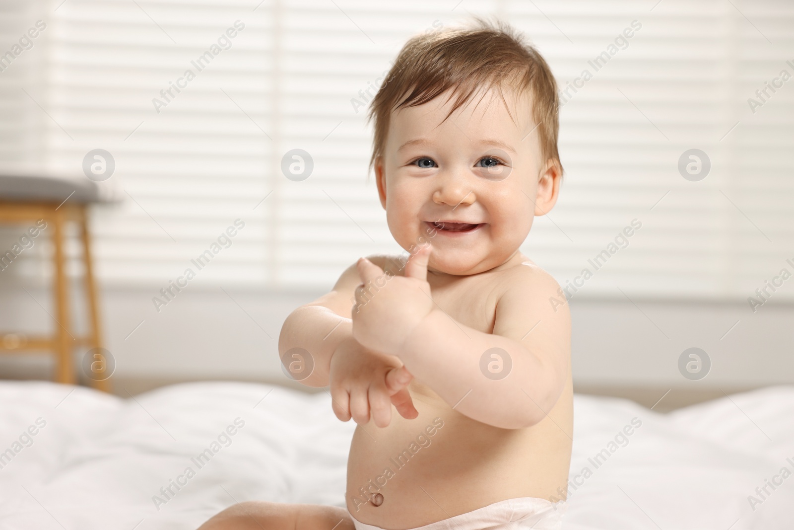 Photo of Cute little baby with moisturizing cream onto body sitting on bed at home