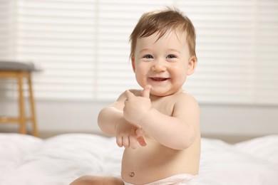 Cute little baby with moisturizing cream onto body sitting on bed at home