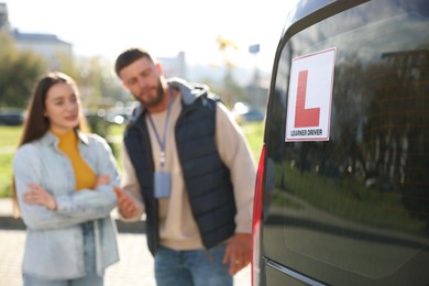 Photo of Learner driver and instructor near car outdoors, selective focus on L-plate. Driving school