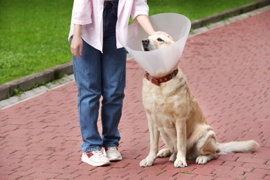 Photo of Woman petting her adorable Labrador Retriever dog in Elizabethan collar outdoors, closeup