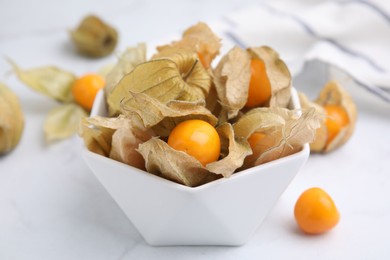 Ripe physalis fruits with calyxes in bowl on white marble table