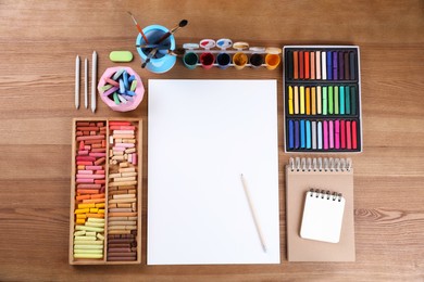 Blank sheet of paper, colorful chalk pastels and other drawing tools on wooden table, flat lay. Modern artist's workplace