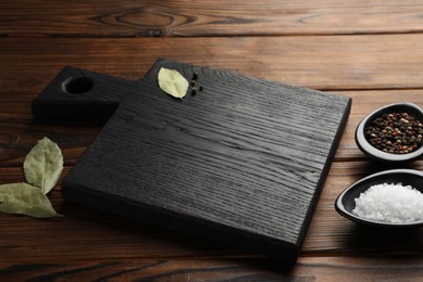 Photo of Black cutting board, salt, pepper and bay leaves on wooden table. Space for text