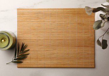 Photo of Bamboo mat, green leaf, vase with plant and burning candle on white marble table, flat lay. Space for text