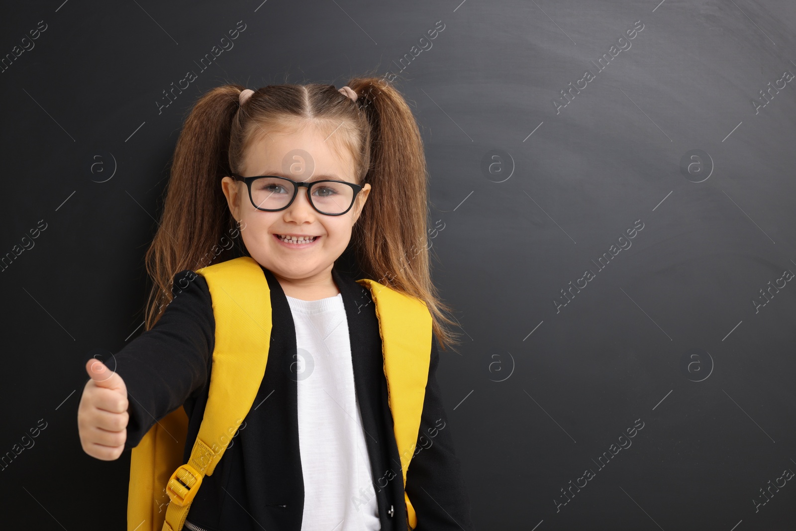 Photo of Happy little school child with backpack showing thumbs up near chalkboard. Space for text