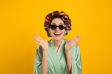 Happy young woman in silk bathrobe with hair curlers and sunglasses on yellow background