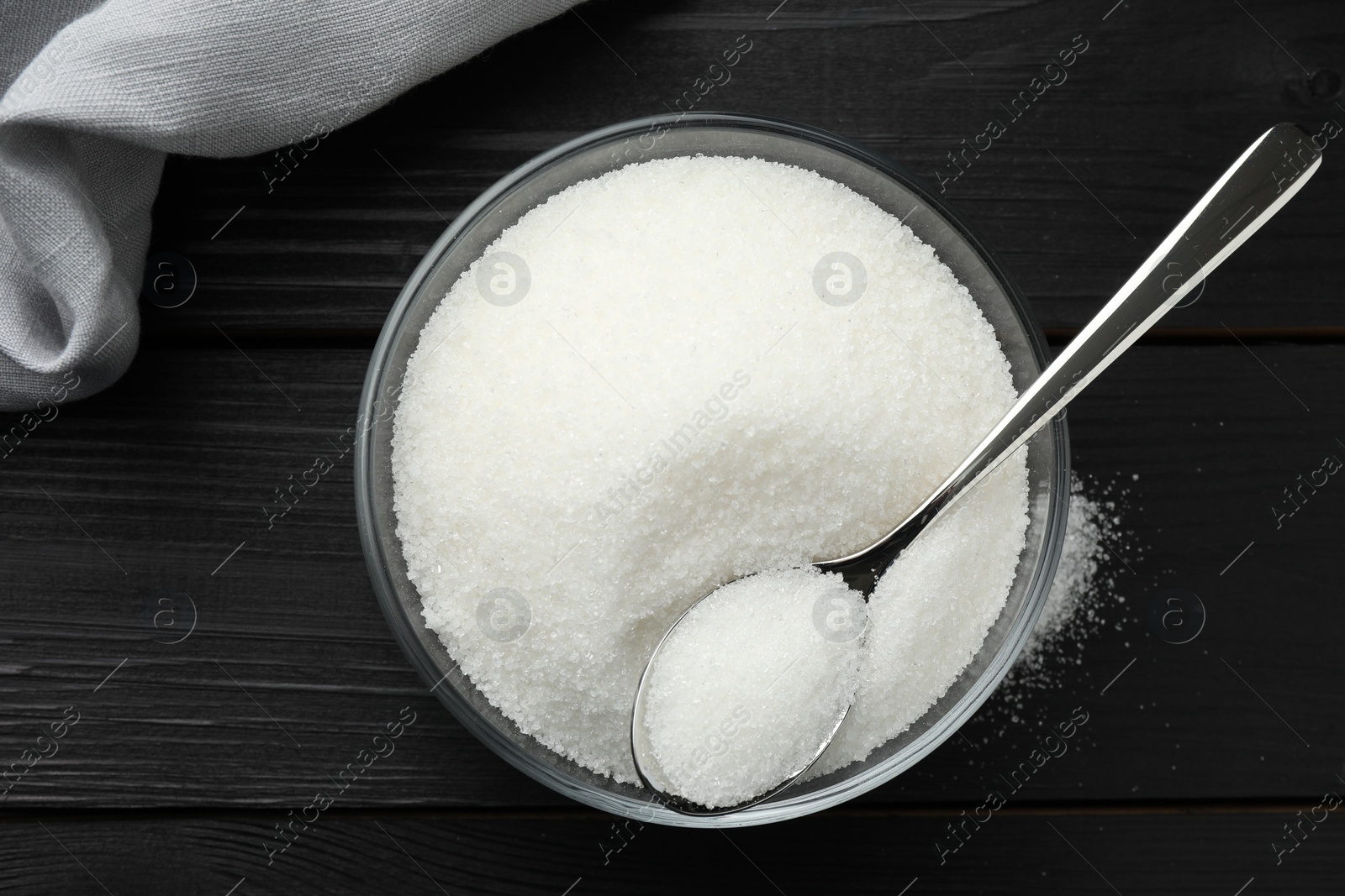 Photo of Granulated sugar in bowl and spoon on black wooden table, top view
