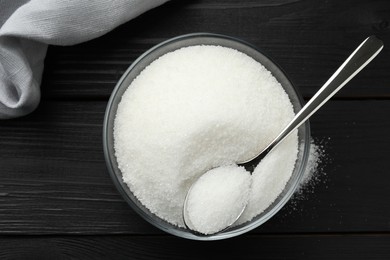 Granulated sugar in bowl and spoon on black wooden table, top view