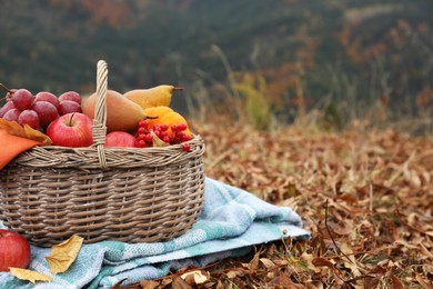 Wicker picnic basket with fruits and plaid on autumn leaves in nature, space for text