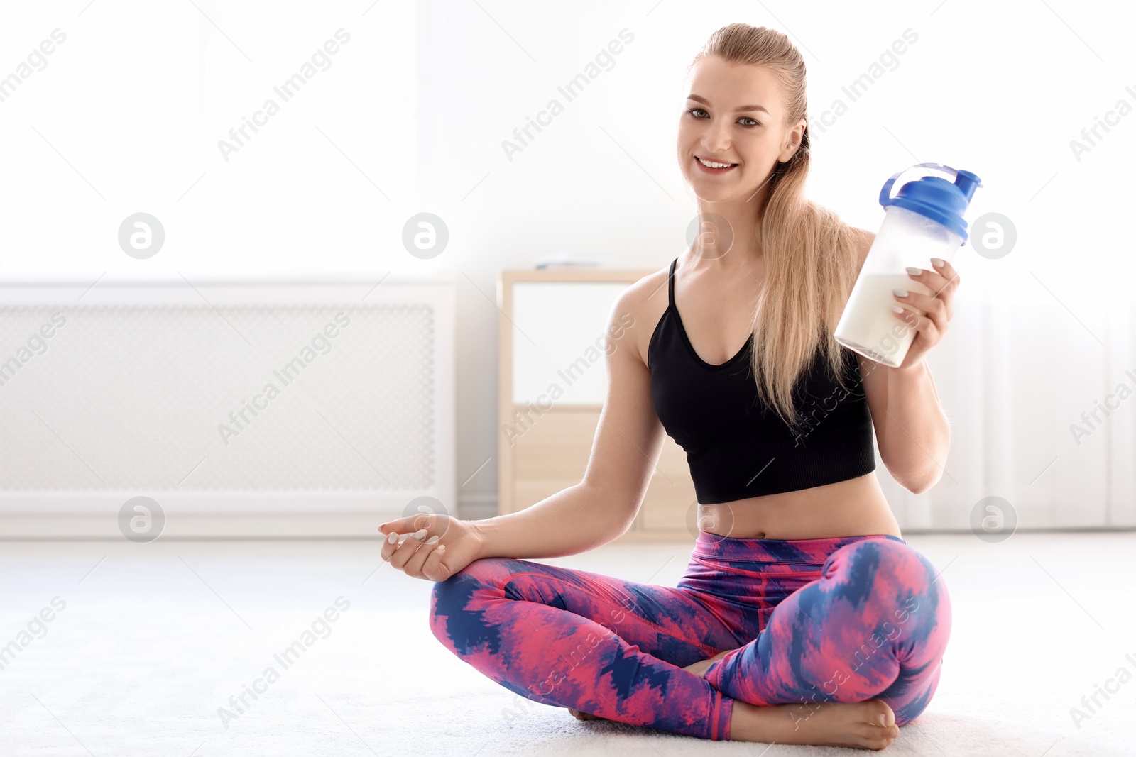 Photo of Young sporty woman with bottle of protein shake sitting on floor at home. Space for text