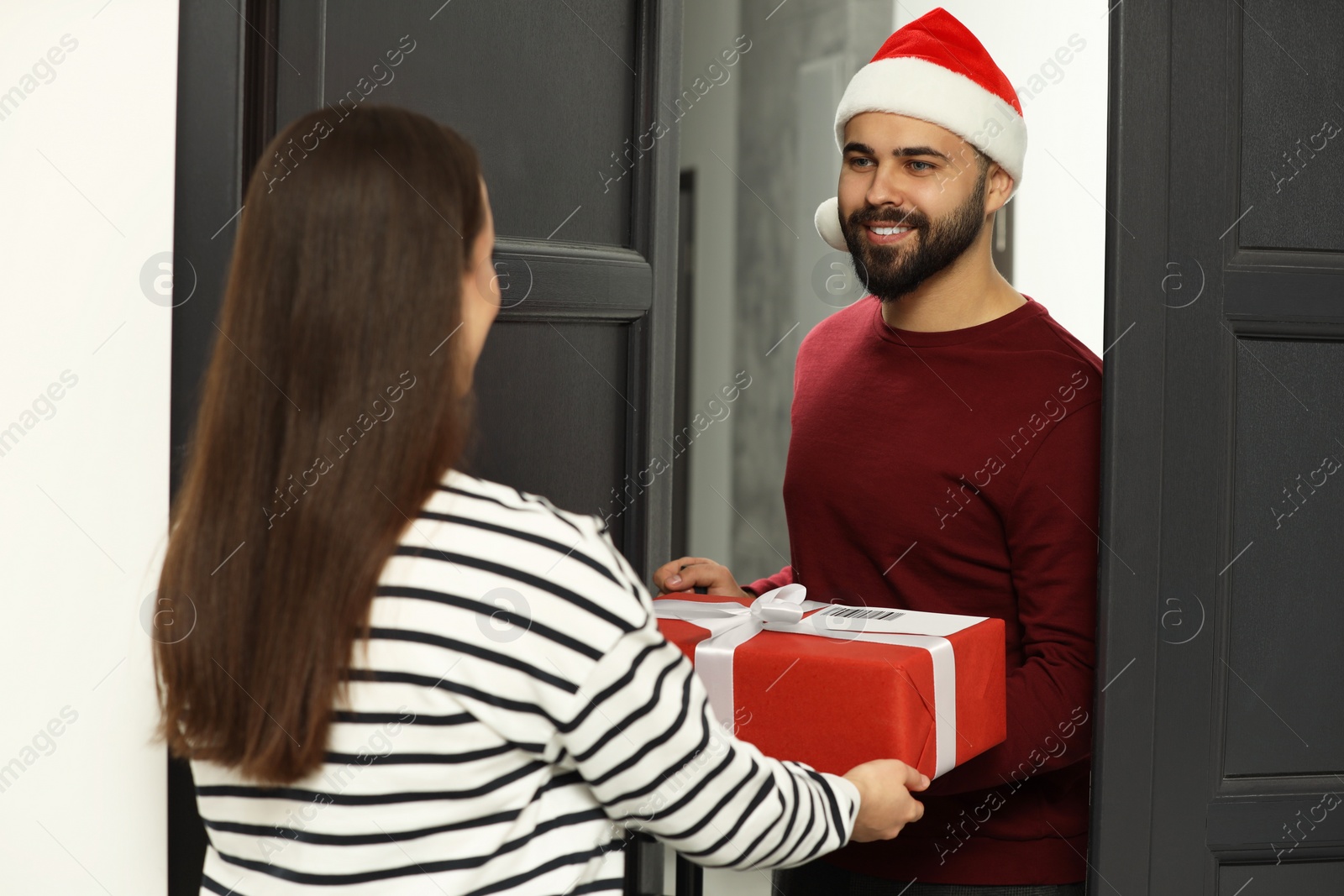 Photo of Courier in Santa hat giving young woman Christmas gift box indoors. Sending present by mail