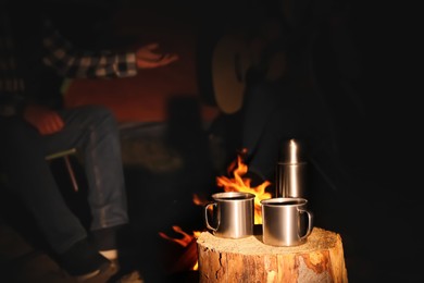 Photo of People near bonfire outdoors at night, focus on stump with metal mugs and thermos. Camping season