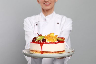 Happy professional confectioner in uniform holding delicious cake on light grey background, closeup
