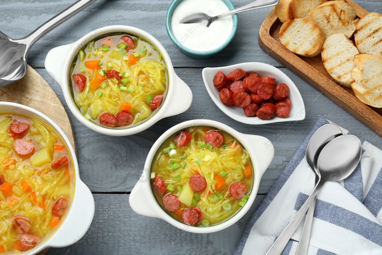 Photo of Delicious sauerkraut soup with smoked sausages and green onion served on grey wooden table, flat lay