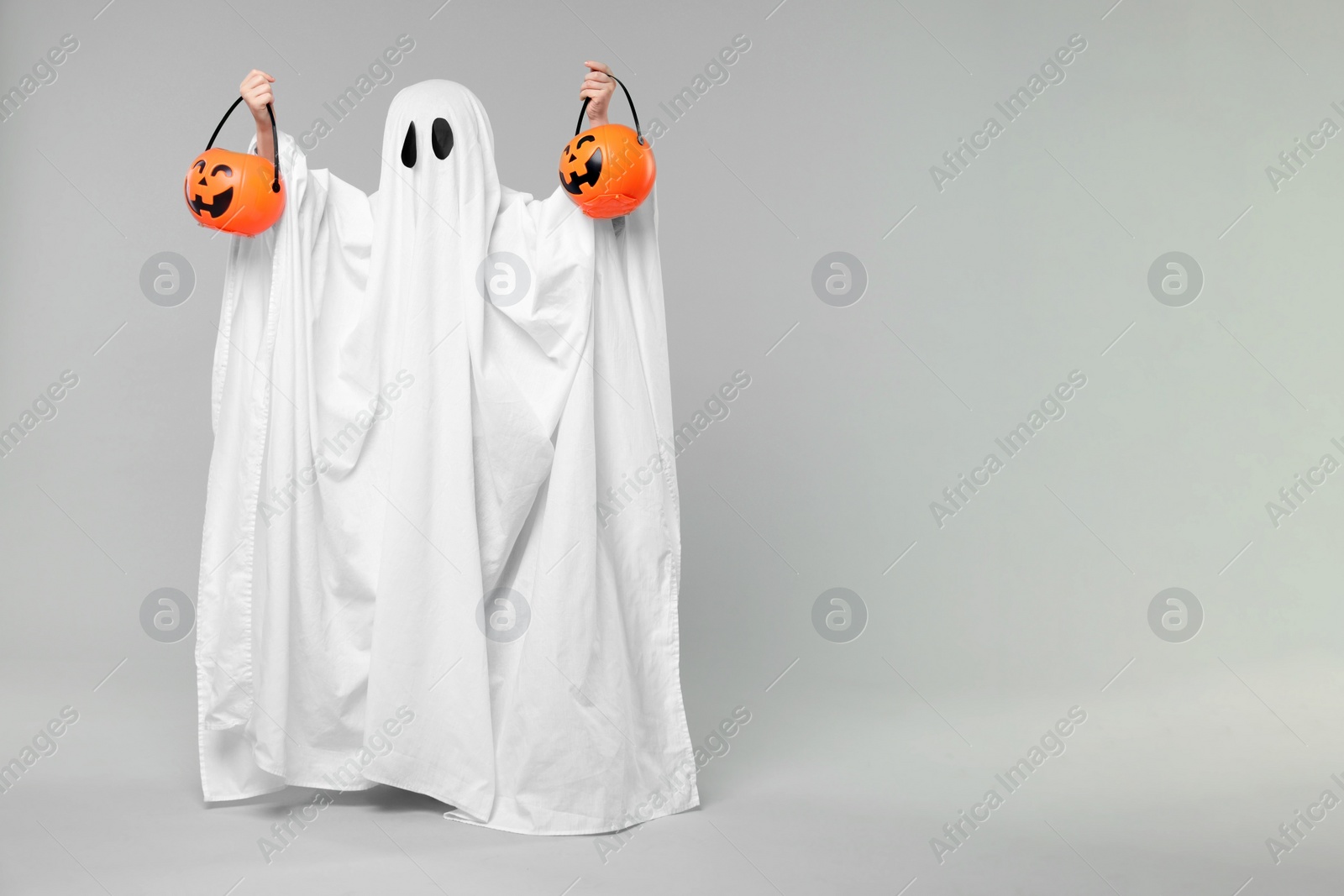 Photo of Child in white ghost costume holding pumpkin buckets on light grey background, space for text. Halloween celebration