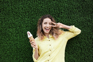 Photo of Happy young woman with delicious ice cream in waffle cone near grass wall