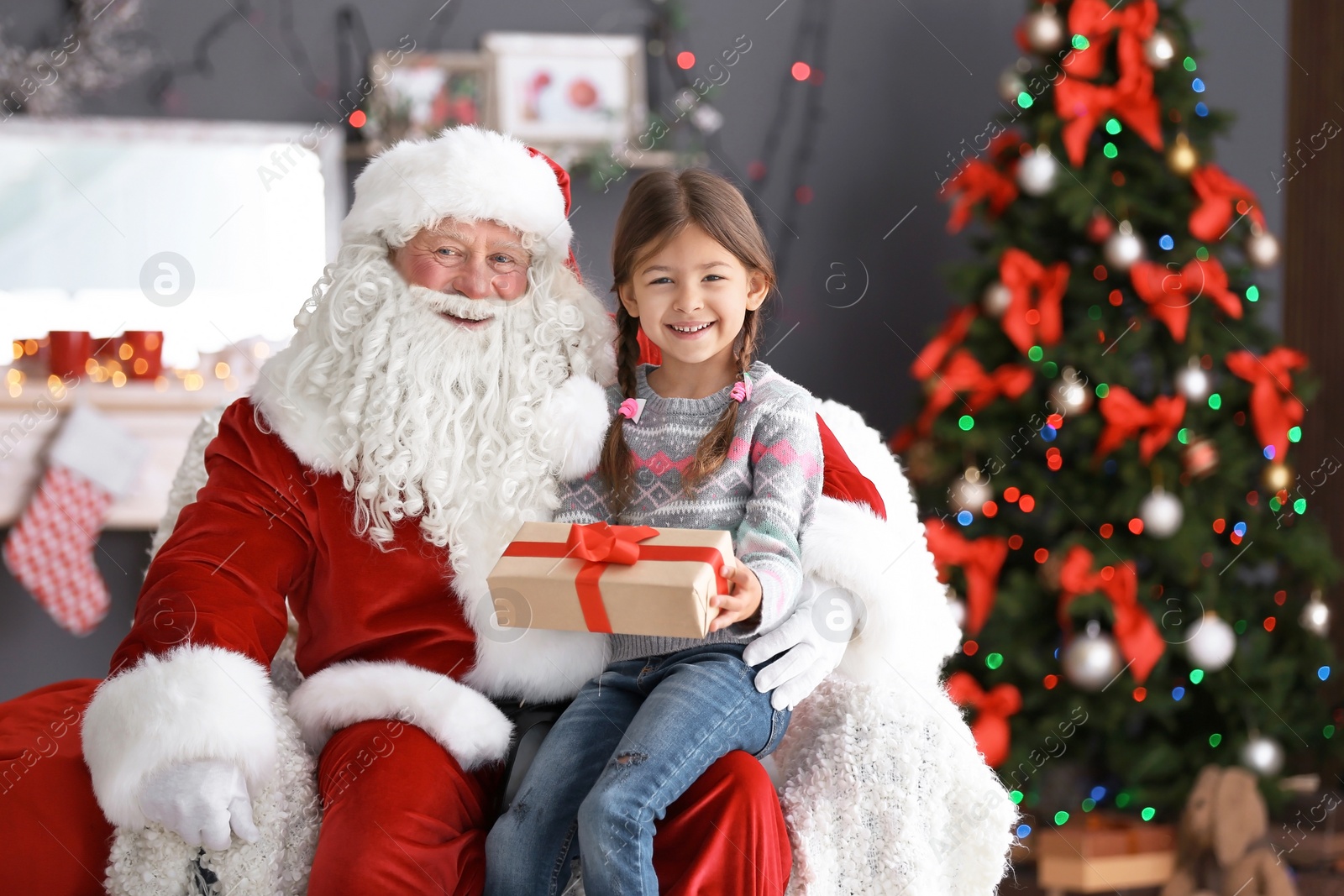 Photo of Little girl with gift box sitting on authentic Santa Claus' lap indoors