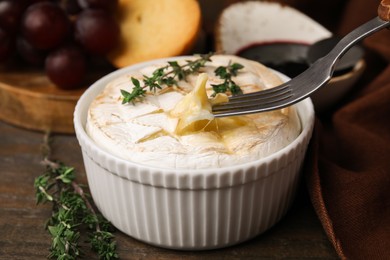 Photo of Eating tasty baked camembert with fork from bowl at wooden table, closeup