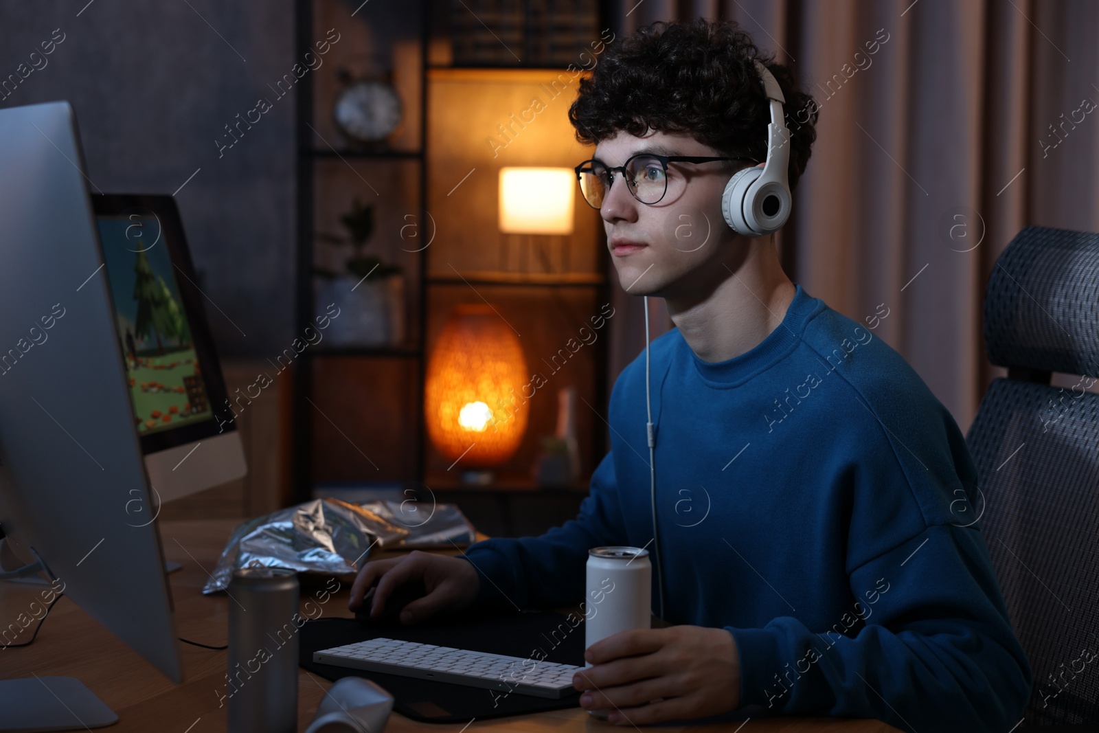 Photo of Young man with energy drink and headphones playing video game at wooden desk indoors