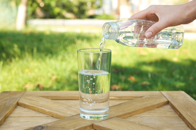 Photo of Woman pouring water from bottle into glass at wooden table outdoors, closeup