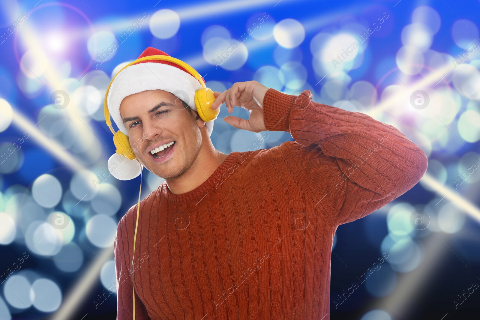 Image of Happy man in Santa hat listening to Christmas music with headphones on bright background, bokeh effect