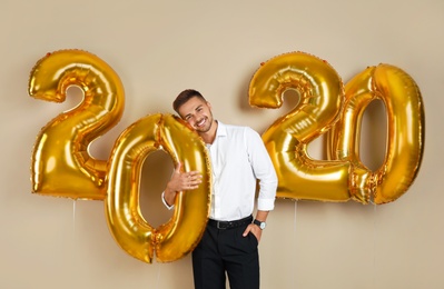 Happy young man with golden 2020 balloons on beige background. New Year celebration