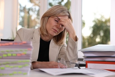 Photo of Overwhelmed woman sitting at table with stacks of documents and folders in office