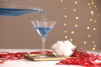 Photo of Cocktail making. Pouring tasty alcoholic drink in glass at white table against beige background with blurred lights, closeup