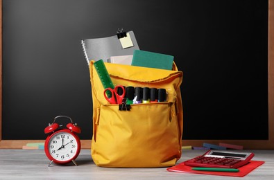 Photo of Backpack with different school stationery on white wooden table near blackboard