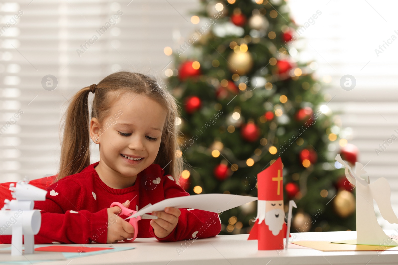 Photo of Cute little girl cutting paper at table with Saint Nicholas toy indoors