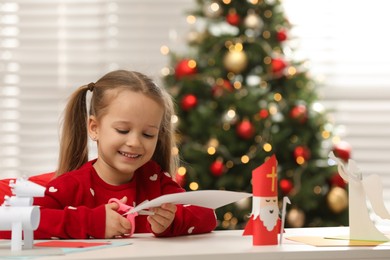 Photo of Cute little girl cutting paper at table with Saint Nicholas toy indoors