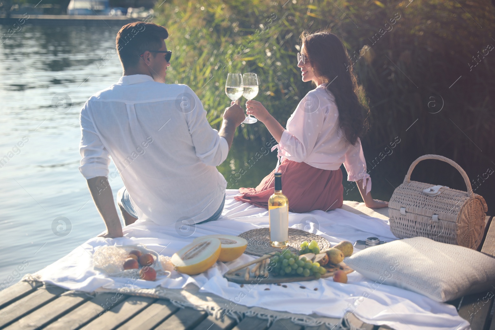 Photo of Couple clinking glasses with wine on pier at picnic, back view