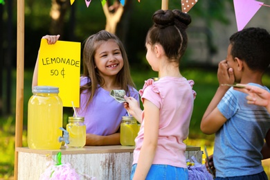 Photo of Little girl selling natural lemonade to kids in park. Summer refreshing drink