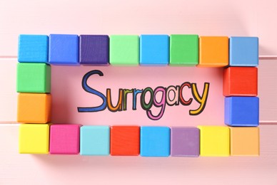 Photo of Word Surrogacy and colorful cubes on pink wooden table, flat lay