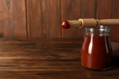 Photo of Tasty barbecue sauce in glass jar and brush on wooden table, closeup. Space for text