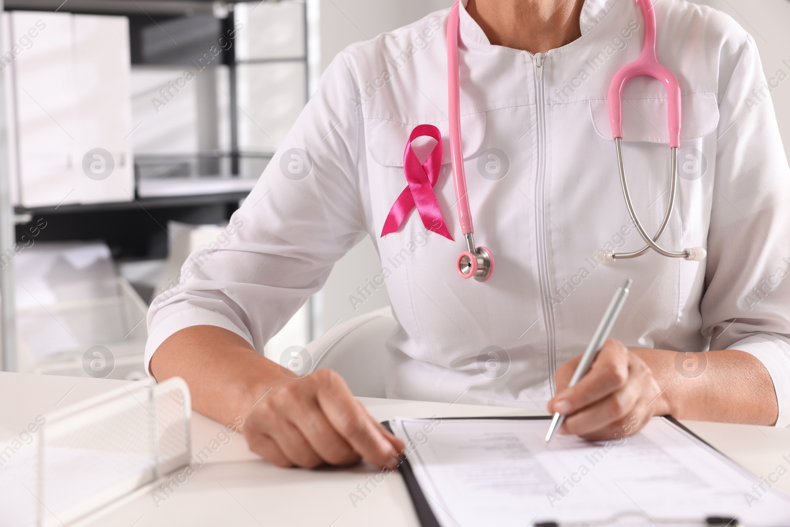 Photo of Doctor with stethoscope and pink ribbon at white desk indoors, closeup. Breast cancer awareness