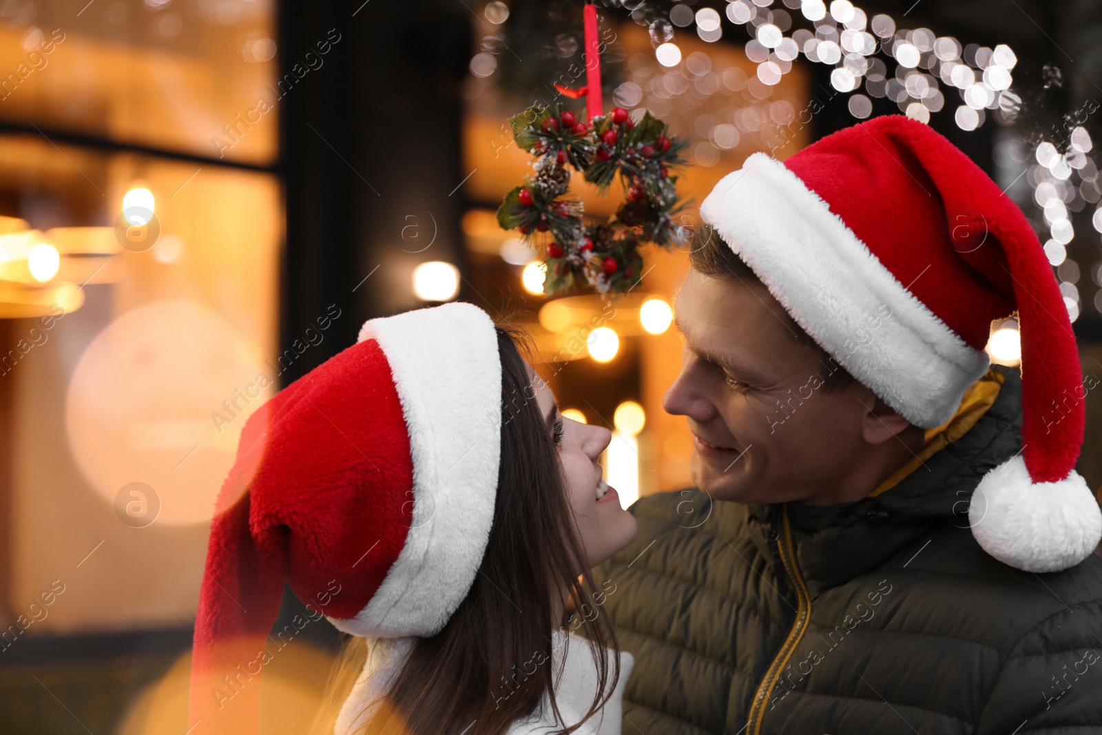 Photo of Happy couple in Santa hats standing under mistletoe wreath outdoors, bokeh effect