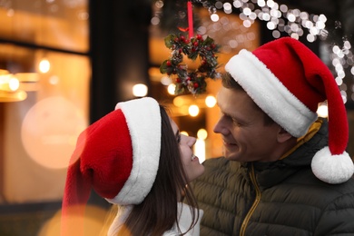 Happy couple in Santa hats standing under mistletoe wreath outdoors, bokeh effect