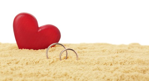 Honeymoon concept. Two golden rings, red wooden heart and sand isolated on white