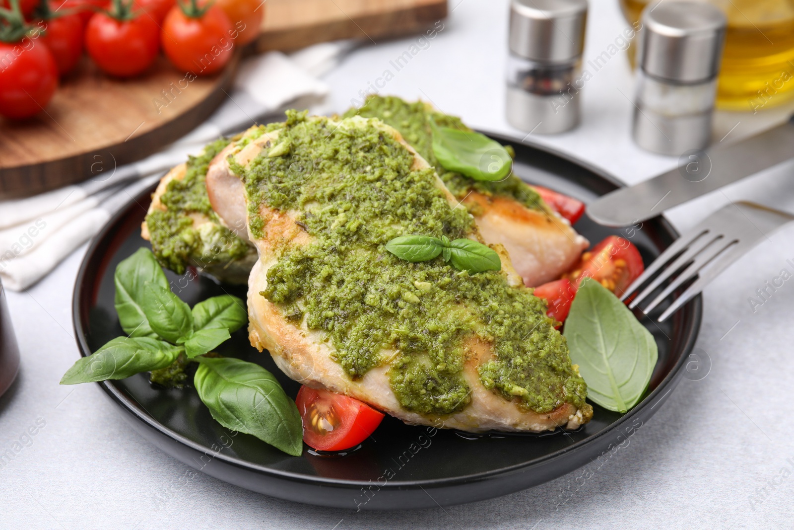 Photo of Delicious chicken breasts with pesto sauce, tomatoes and cutlery served on light table, closeup