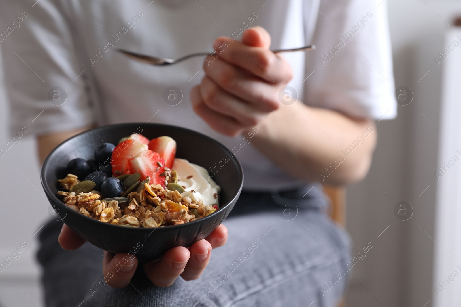 Photo of Woman eating tasty granola with berries, yogurt and seeds indoors, closeup