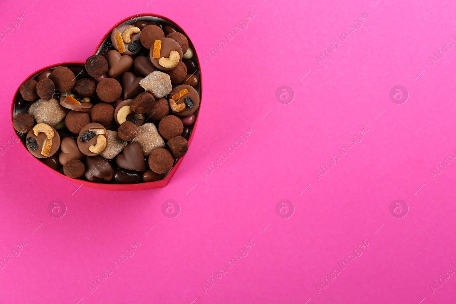 Photo of Different delicious chocolate candies in heart shaped box on pink background, top view. Space for text