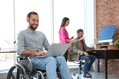 Photo of Young man in wheelchair with laptop and his colleagues at office