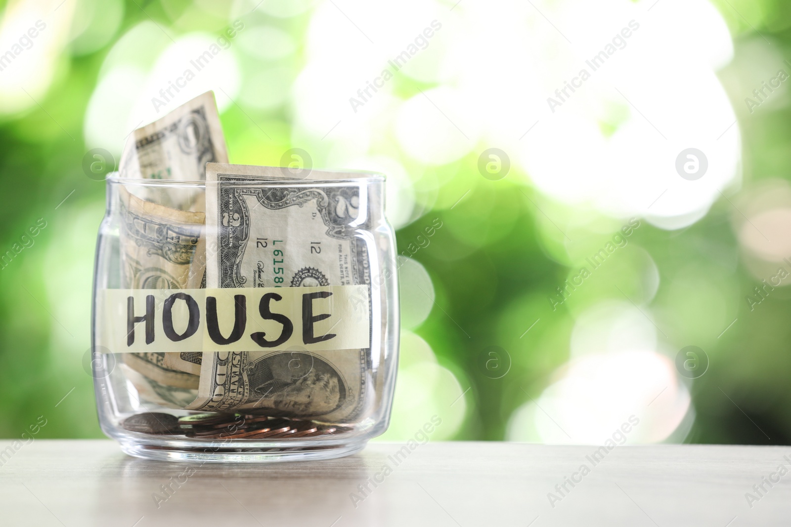 Photo of Glass jar with money and label HOUSE on table against blurred background. Space for text