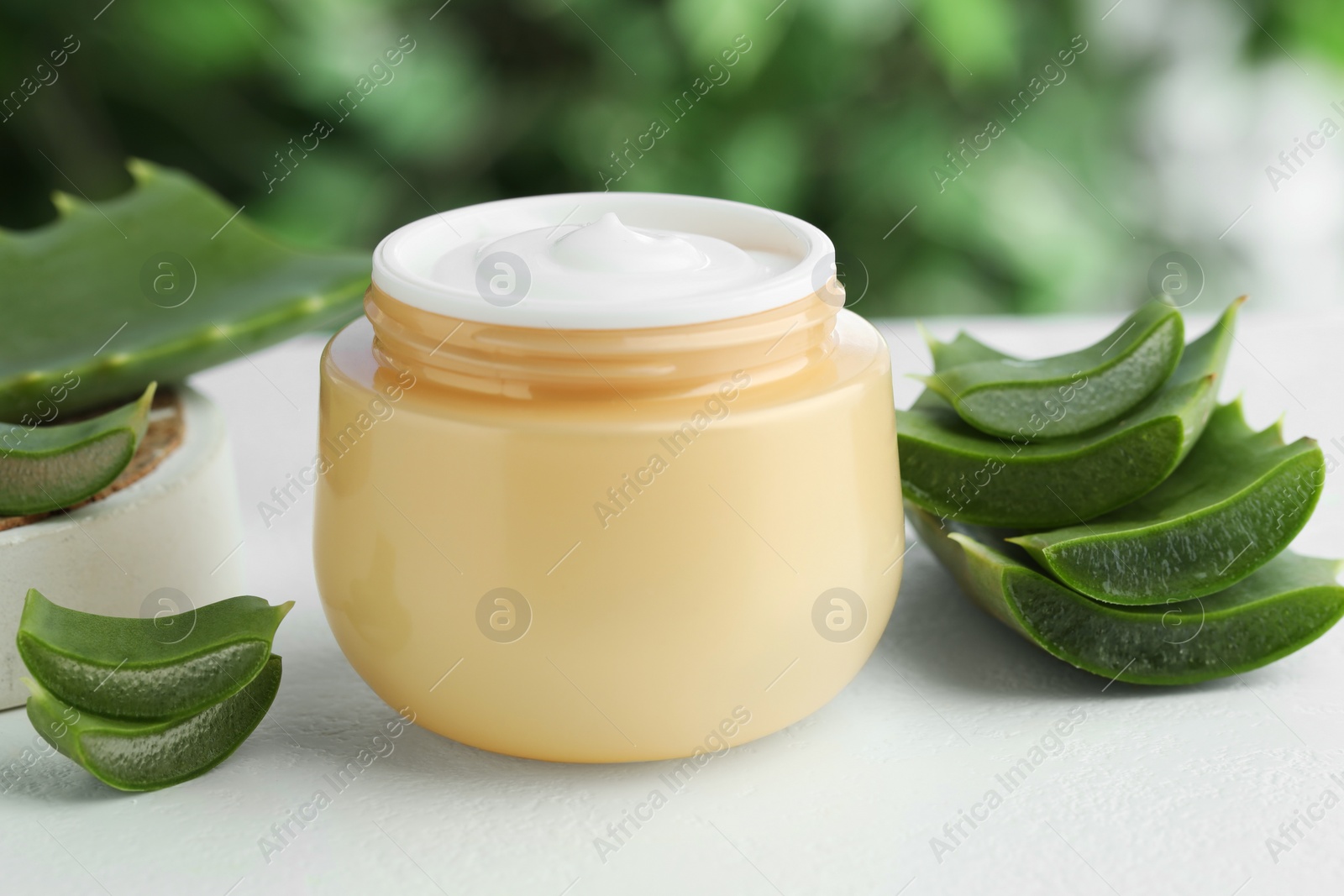 Photo of Jar with cream and cut aloe leaf on white table against blurred green background, closeup