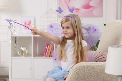 Photo of Cute little girl in fairy costume with violet wings and magic wand in armchair at home