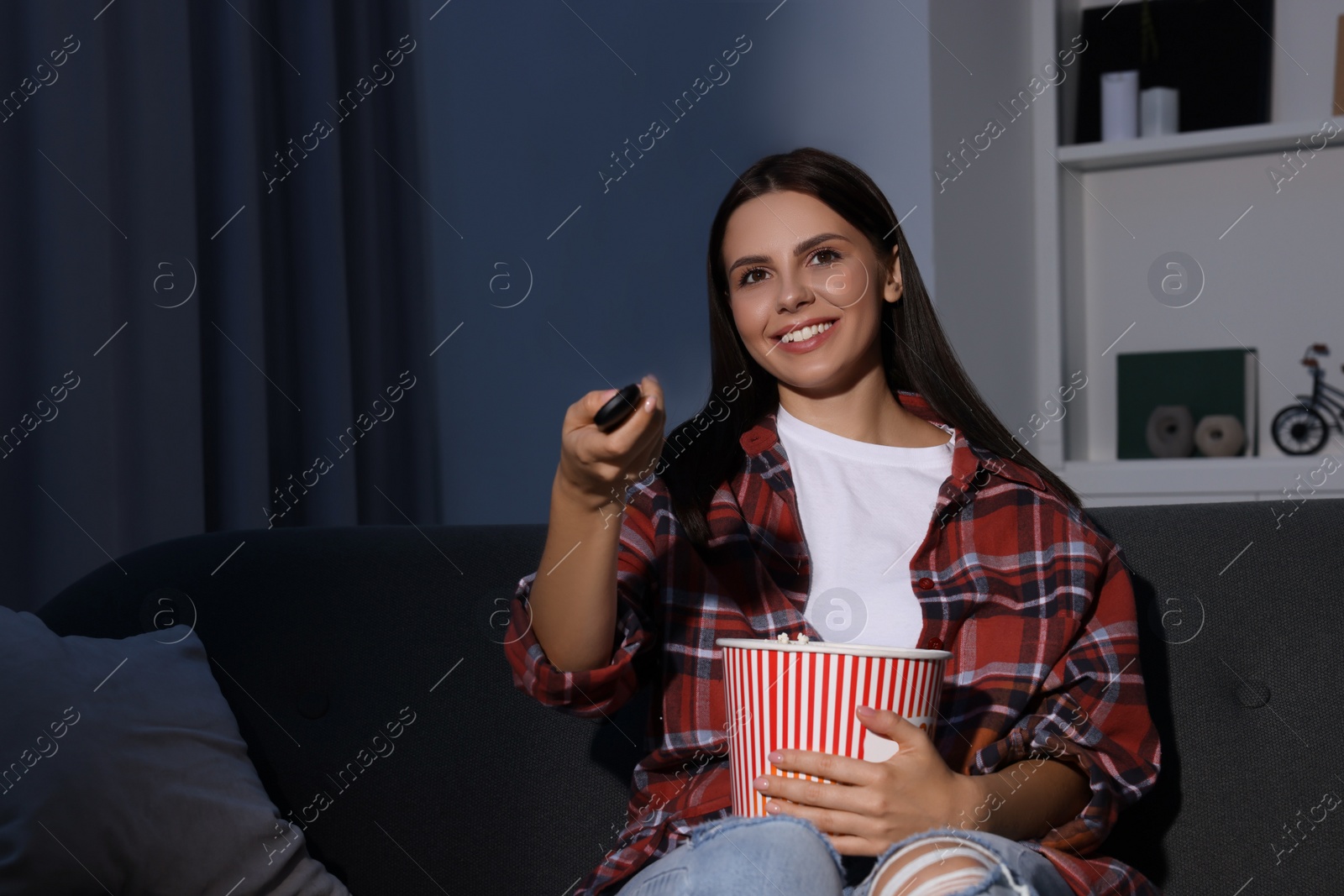 Photo of Happy woman holding popcorn bucket and changing TV channels with remote control at home in evening